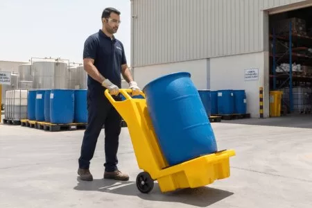 Worker transporting a blue chemical drum using a yellow drum handling trolley in an industrial yard.